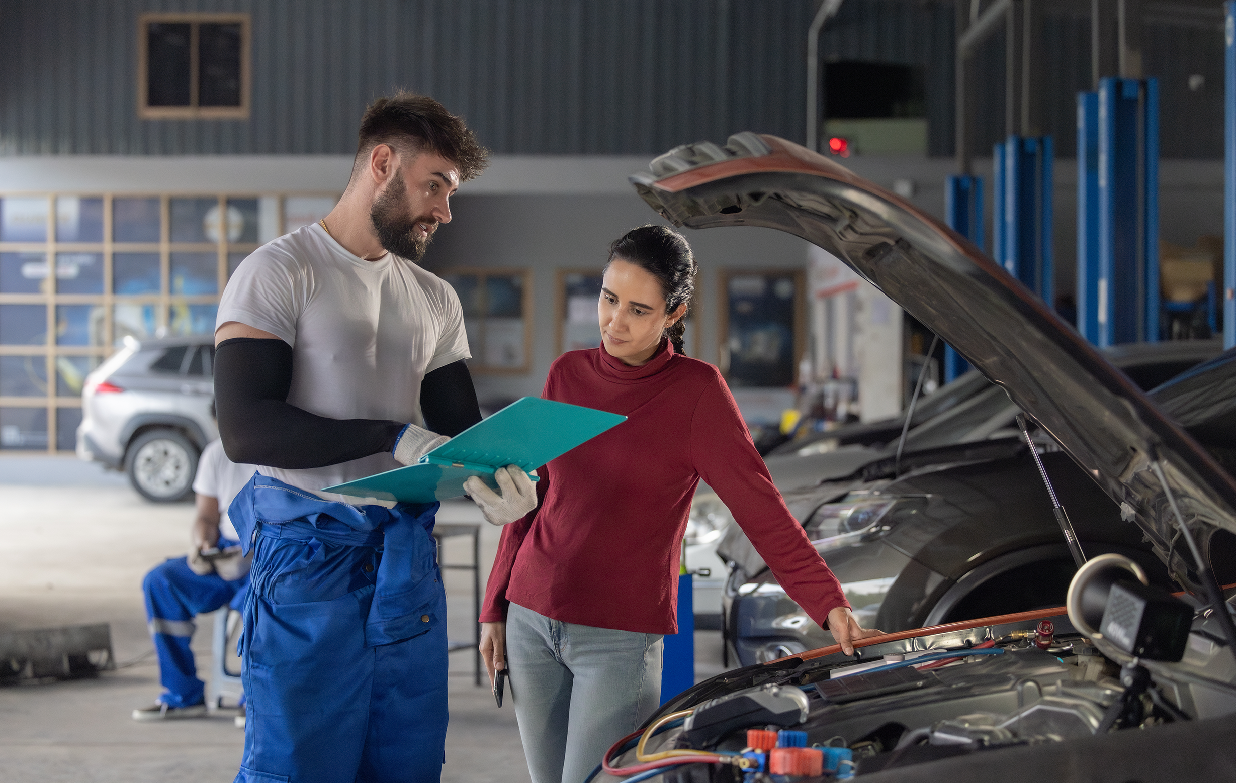 car mechanic and customer looking at clip board with open car hood in a car garage