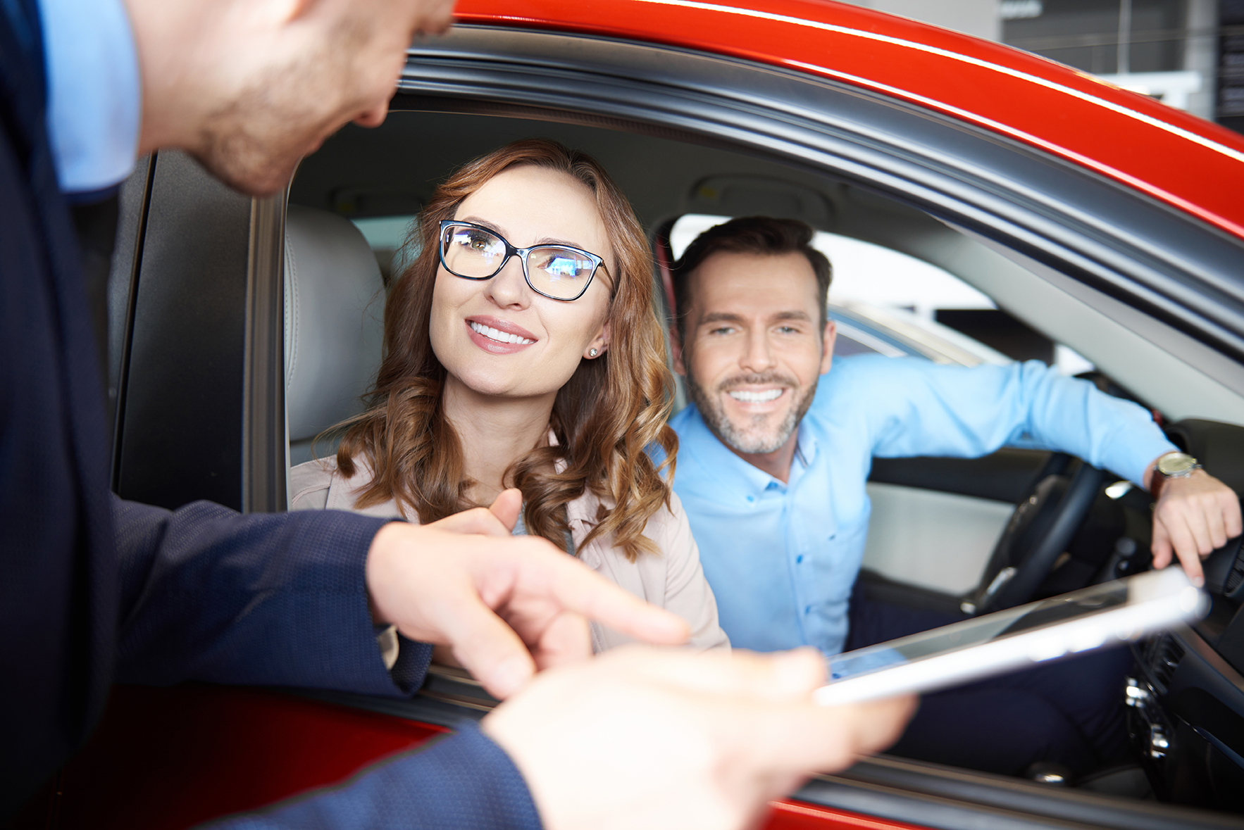 couple sitting in red car with a person talking to them with a tablet through the window