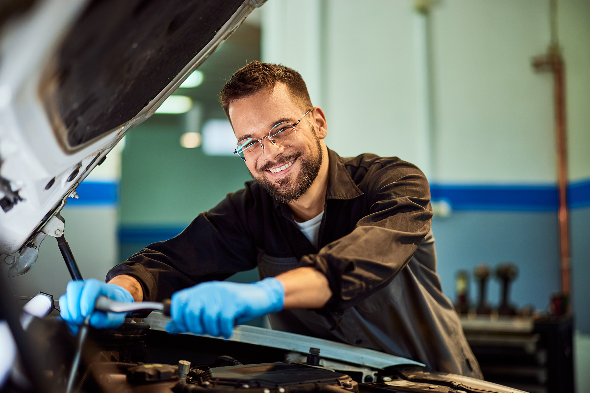 mechanic working on a car with glasses adn blue gloves with a smile in a garage under the hood of the car
